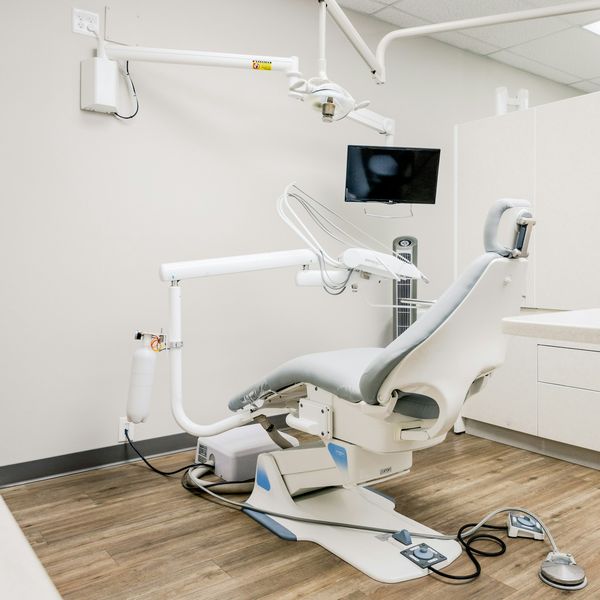 A bright treatment room: one dental chair beside a window, white cabinets and a screen on the wall