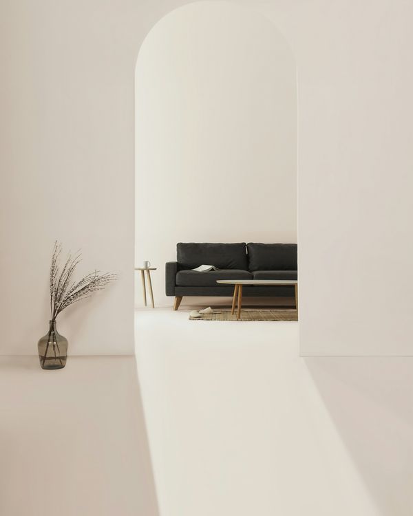 A quiet cream room seen through an arch: a low black sofa and a plant in a stone pot